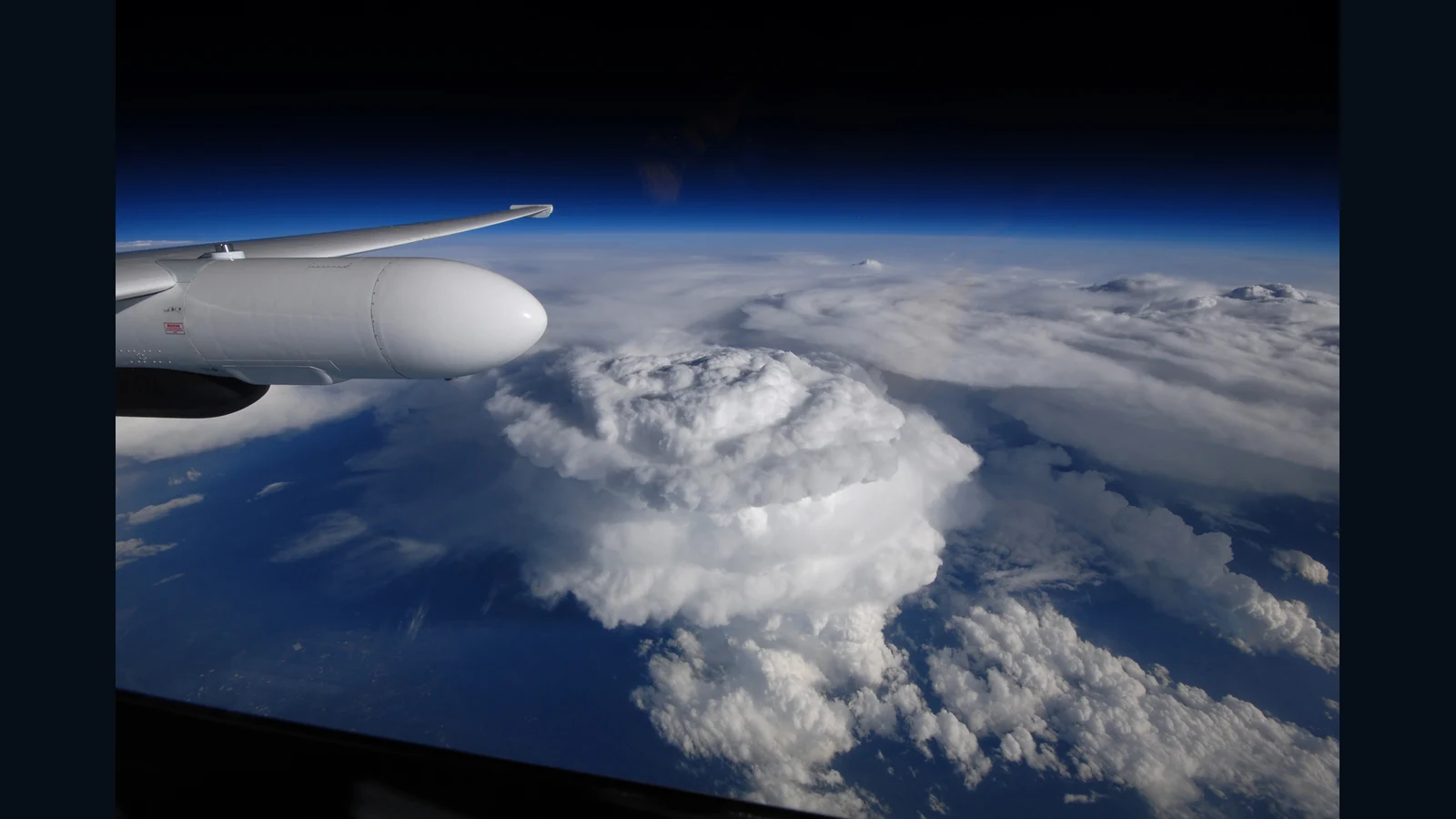 ER-2研究機の窓から見下ろす雷雲。手前に機体の一部、雲の上面に盛り上がる頂部、奥に濃い青の空が見える